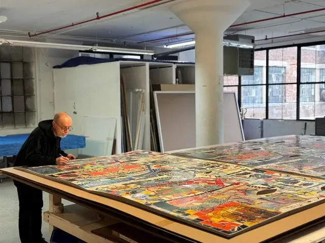 A printmaker leaning over a large, densely coloured screenprint laid flat on a table in a loft studio, with racks of panels behind and windows along the far wall.